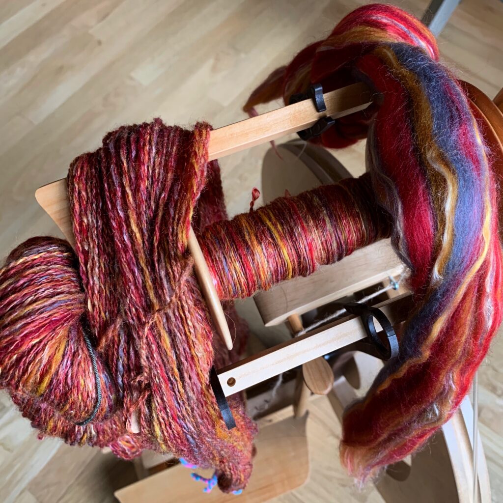 A top-down view showing the progression from fiber to singles to 2-ply yarn, with the yarn & fiber draped over a castle-style spinning wheel whose bobbin is half-full of singles The fiber/singles/yarn is mostly red with hints of yellow/gold, grey, and blue.