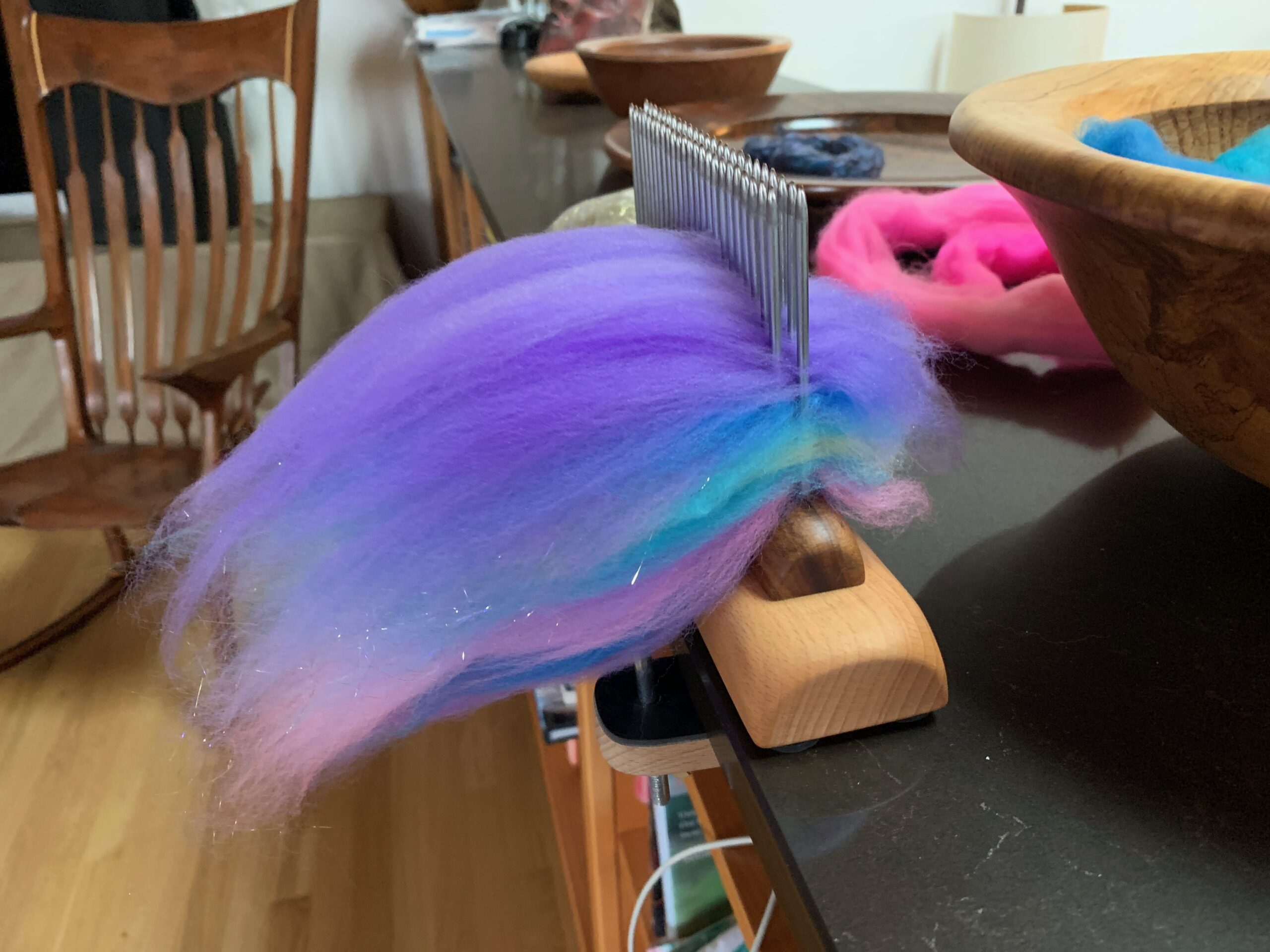 A blending comb clamped onto a counter, with layers of baby pink, blue, yellow, and purple fiber on it, along with some angelina stellina sparkles. In the background you can see more fiber, several wooden bowls my partner made, and a gorgeous wooden rocking chair. The fiber is from Frabjous Fibers, one of their Three Feet of Fiber bags.
