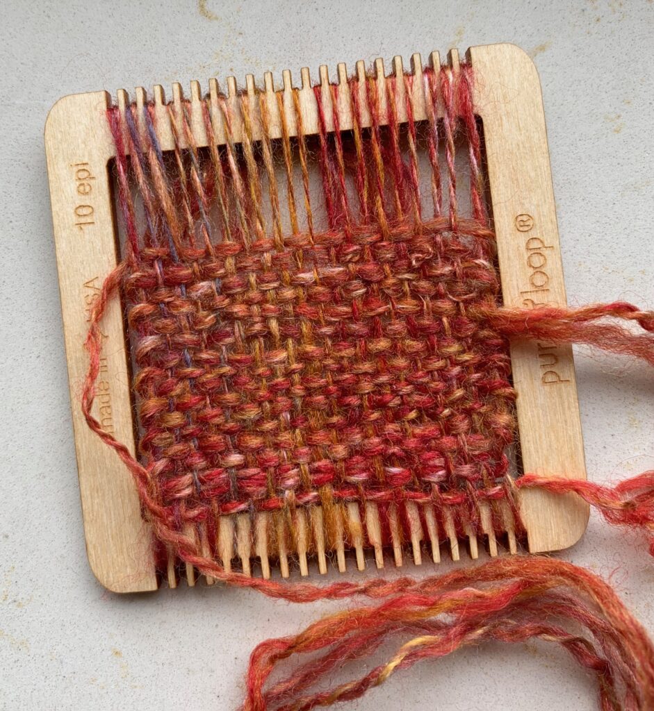 The same yarn as in the previous image, in shades of mostly red, yellow, and white, now woven on a tiny weaving frame. It's about 3/4 complete, and the weave is fairly tight. The frame is from purl&loop, and it's a 10 ends per inch (EPI) size.