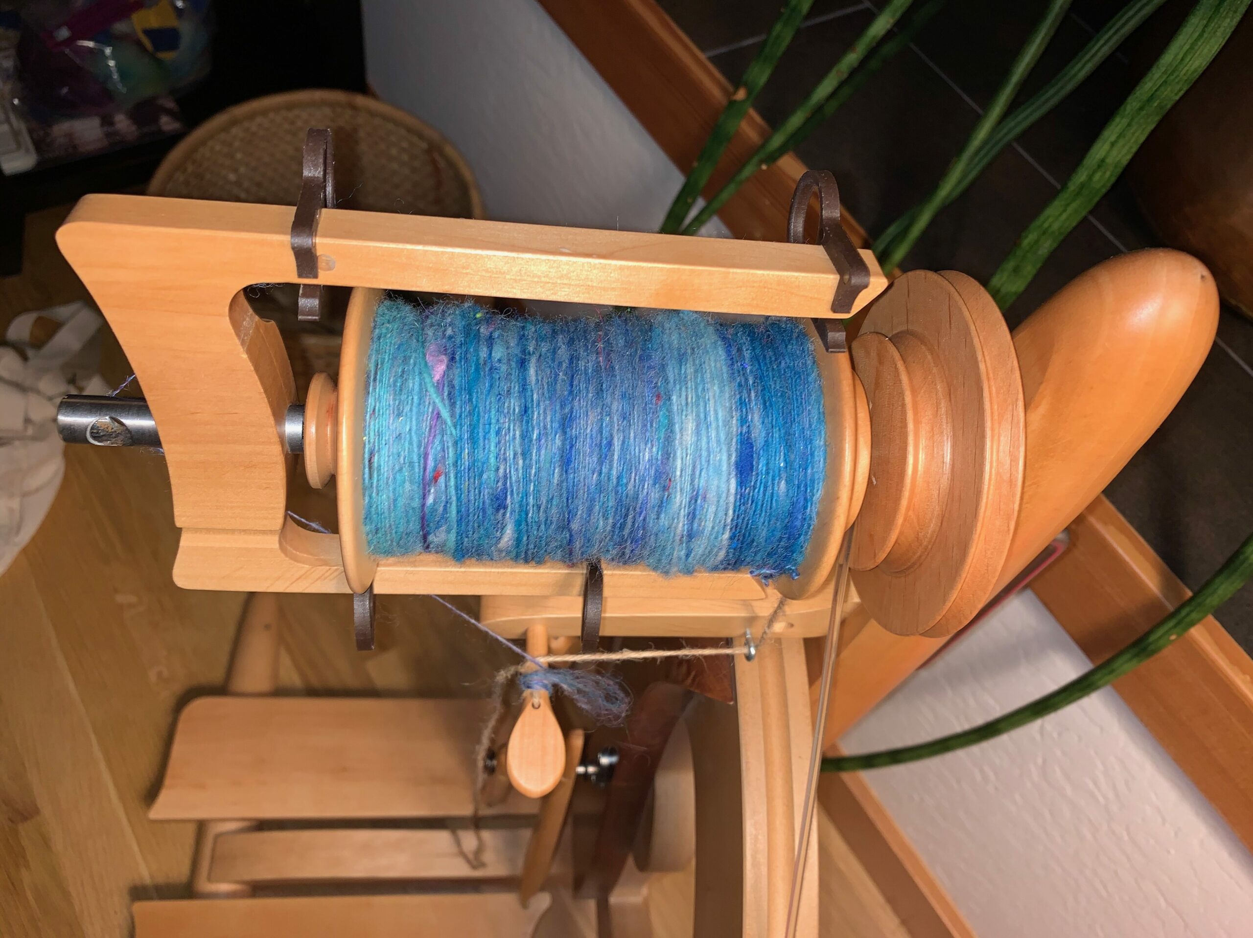Nearly top-down view of a castle-style spinning wheel with a bobbin mostly full of finely spun singles. The yarn is in shades of blue with hints of purple.