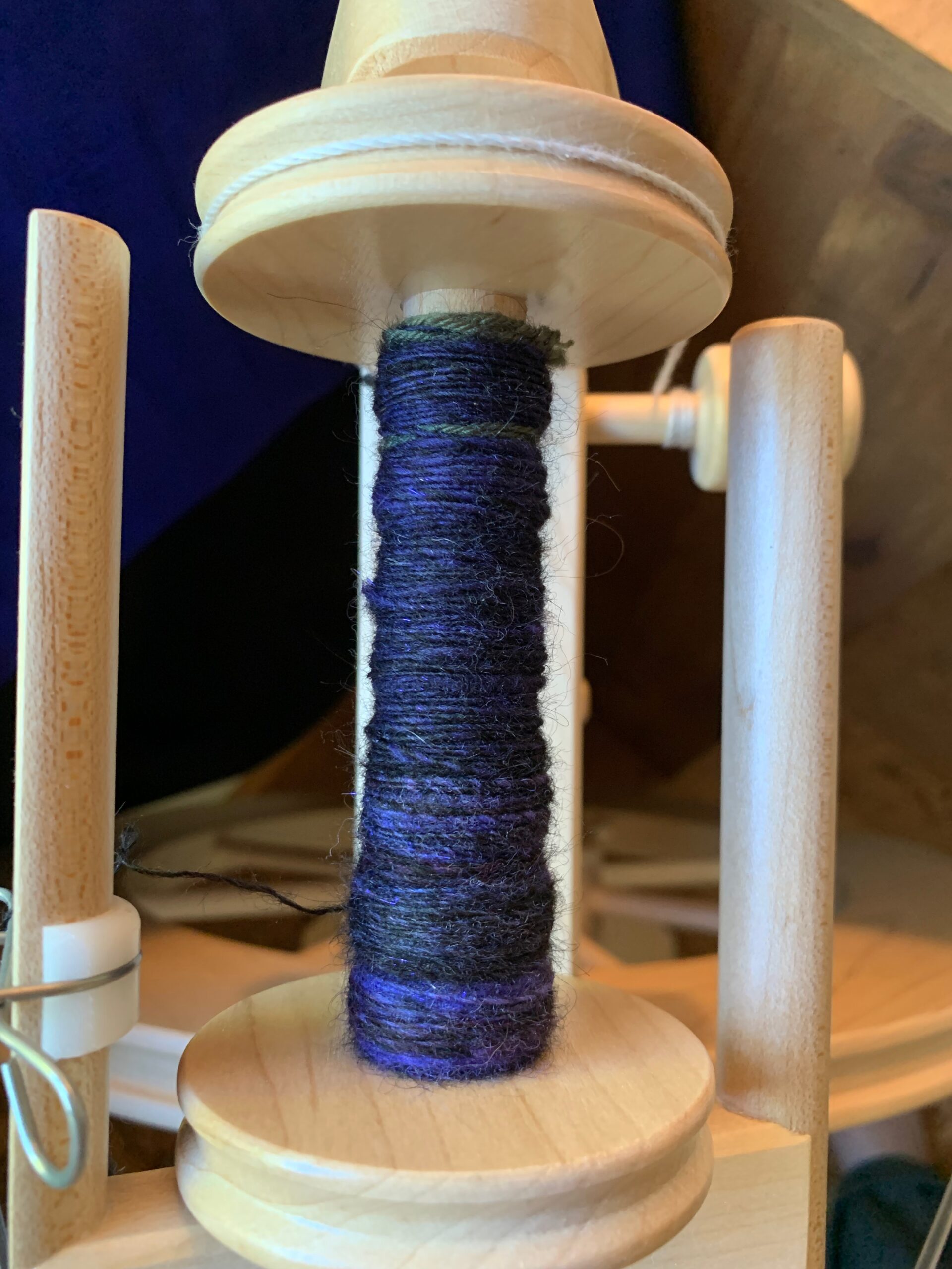 Top-down view of the bobbin and flyer of a spinning wheel, with a blend of black and dark purple singles wound on it, and some green yarn that is the leader that attaches the new yarn to the bobbin.