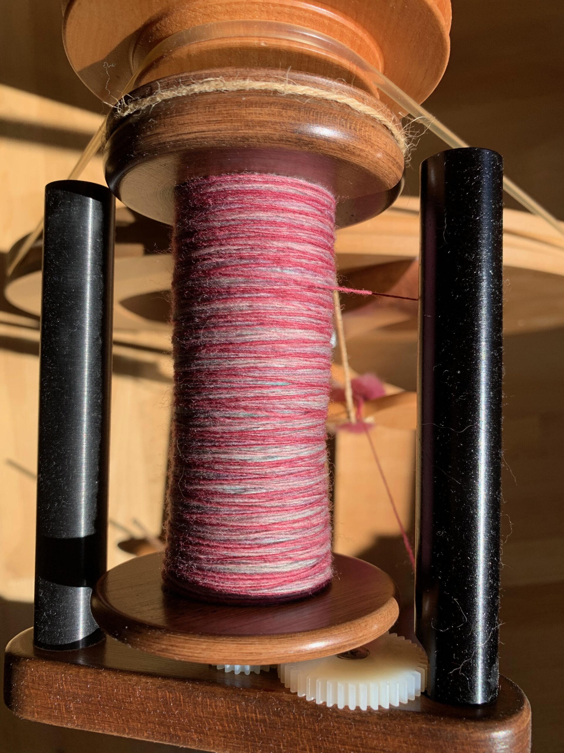 Top-down view of a bobbin on a spinning wheel with a Woolee Winder flyer. The bobbin has a singles (unplied yarn) in shades of magenta and pale green. The magenta is dominant.