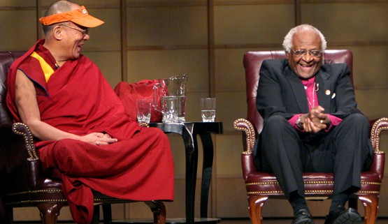 The Dalai Lama and Desmond Tutu laughing & having fun together. They're on a stage in leather armchairs, with a table with glasses of water nearby. The Dalai Lama is laughing and looking at Reverend Tutu, while sitting with his legs criss-crossed and hands together in his lap. Desmond Tutu has his hands clasped between his knees and is laughing with his eyes closed.